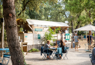 Udendørs caféområde i Huttopia Oléron les Pins feriepark, med folk der nyder mad og sollys blandt træer.