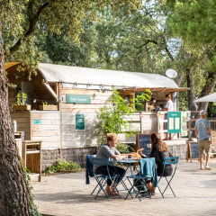Zona de cafetería al aire libre en Huttopia Oléron les Pins, con personas comiendo entre árboles y naturaleza.