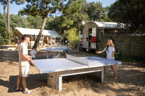 Personas juegan tenis de mesa al aire libre en un parque de vacaciones boscoso con casita y tobogán al fondo.