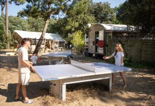 Des personnes jouent au tennis de table en plein air dans un parc de vacances boisé, avec cabane et toboggan.