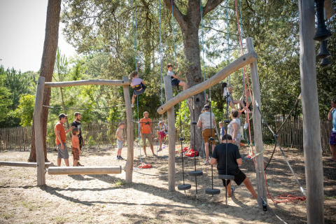 Niños y adultos disfrutan de una zona de juegos de escalada al aire libre bajo los árboles en Huttopia Oléron les Pins.