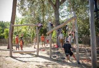 Børn og voksne leger på en udendørs klatrelegeplads i skyggen af træer ved Huttopia Oléron les Pins.