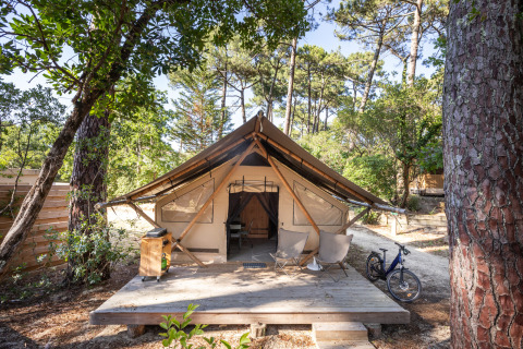 Trapper safari tent with wooden deck, two chairs, and a bicycle, surrounded by trees in sunlight.