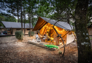 Famiglia cena davanti a una tenda safari Trapper illuminata presso Huttopia Arcachon, Francia, tra gli alberi.