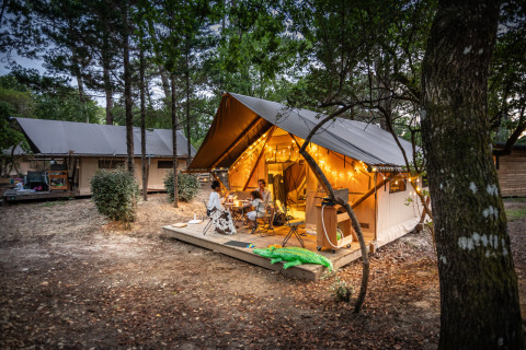 Famiglia cena davanti a una tenda safari Trapper illuminata presso Huttopia Arcachon, Francia, tra gli alberi.