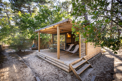 Træchalet Evasion i skoven med overdækket terrasse, borde, stole og afslappende liggestole på sandjord.