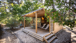 Chalet Evasion aus Holz im Wald mit überdachter Terrasse, Sitzgruppe und Liegestühlen auf Sandboden.