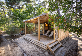 Chalet Evasion aus Holz im Wald mit überdachter Terrasse, Sitzgruppe und Liegestühlen auf Sandboden.