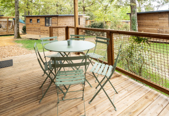 Udendørs træterrasse med bord og stole ved Chalet Evasion, Huttopia Arcachon, omgivet af skov og hytter.