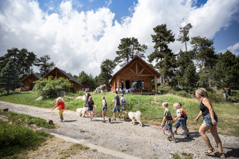 Familias y niños pasean perros frente a cabañas en el parque vacacional Huttopia Font-Romeu, Occitania, Francia.