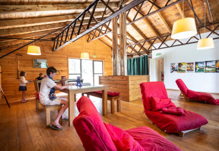 Sala relax in legno con bambini e poltrone rosse presso Huttopia Font-Romeu, Occitania, Francia.