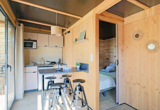 Lichte, moderne keuken met barkrukken en zicht op slaapkamer in Chalet Victoria, Huttopia Arcachon, Frankrijk.