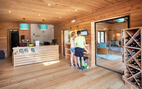 Reception area at Huttopia Lac d’Aiguebelette holiday park with guests, wooden decor, and wine rack.