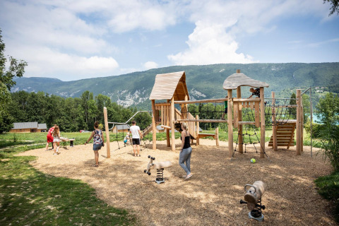 Parque infantil de madera en Huttopia Lac d’Aiguebelette con niños y montañas al fondo, en Francia.