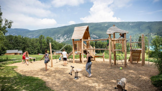 Parque infantil de madera en Huttopia Lac d’Aiguebelette con niños y montañas al fondo, en Francia.