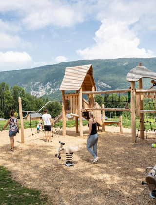 Parque infantil de madera en Huttopia Lac d’Aiguebelette con niños y montañas al fondo, en Francia.