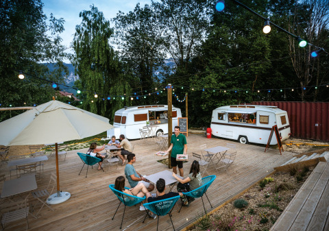 Zona de comedor al aire libre con caravanas, luces y visitantes en Huttopia Lac d’Aiguebelette, Francia.