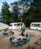 Zona de comedor al aire libre con caravanas, luces y visitantes en Huttopia Lac d’Aiguebelette, Francia.