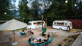 Zona de comedor al aire libre con caravanas, luces y visitantes en Huttopia Lac d’Aiguebelette, Francia.