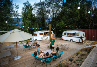 Buitenterras met caravans, lichtslingers en vakantiegangers bij Huttopia Lac d’Aiguebelette in Frankrijk.