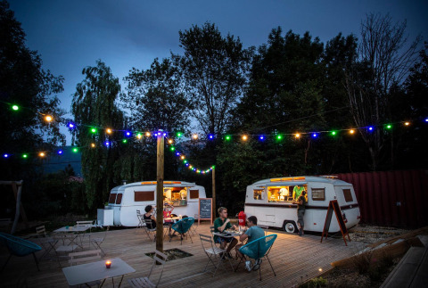 Evening at Huttopia Lac d’Aiguebelette with food trucks, string lights, and people dining on an outdoor deck.