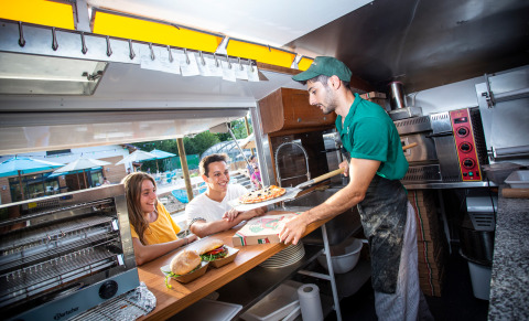 Un chef de pizza sirve una pizza recién hecha a dos jóvenes sonrientes en una food truck en Huttopia Lac d’Aiguebelette, Francia.