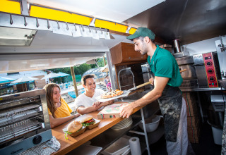 Een pizzachef serveert verse pizza aan twee lachende jongeren bij een foodtruck in Huttopia Lac d’Aiguebelette, Frankrijk.