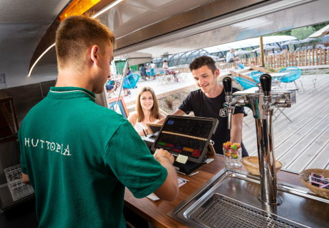 Due ospiti ordinano da bere a un barista con maglietta verde Huttopia in un villaggio vacanze in Francia.