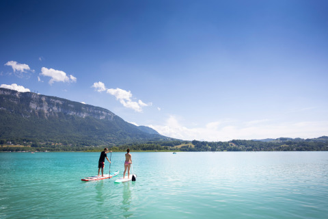 Dos personas hacen paddle surf en un lago turquesa con montañas al fondo en Huttopia Lac d’Aiguebelette.