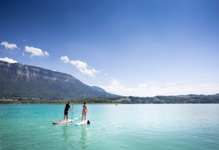 Twee mensen staan op een paddleboard op een helder meer met bergen bij Huttopia Lac d’Aiguebelette.