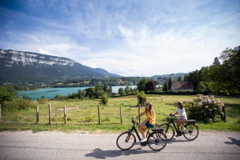 Dos personas montan en bicicleta por una carretera con vistas al lago y montañas en Huttopia Lac d’Aiguebelette, Francia.