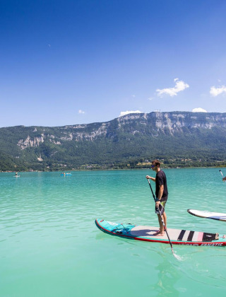 Personas practicando paddle surf en un lago turquesa cerca de Saint Alban de Montbel, rodeados de montañas.
