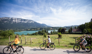 Tres personas pasean en bicicleta por un camino rural con vistas a lago, montañas y prados cerca de Saint Alban de Montbel.