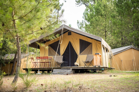 Tienda safari Toile&Bois tent Sweet en el bosque, con terraza y sillas, rodeada de árboles verdes.
