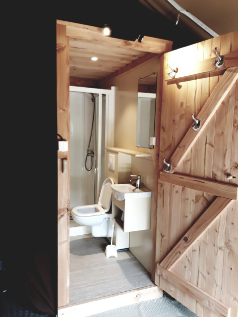 Vista interior de un baño en tienda safari con puerta de madera, inodoro, lavabo y cabina de ducha.