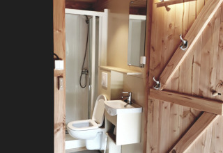 Vista interior de un baño en tienda safari con puerta de madera, inodoro, lavabo y cabina de ducha.