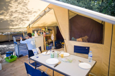 Vista interna di una tenda safari con tavolo, cucina e una persona a Huttopia Oléron les Pins, Francia.