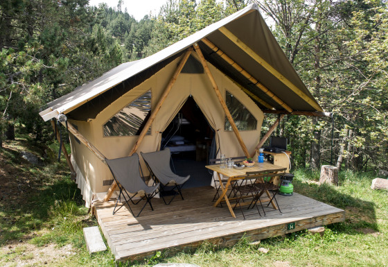 En canadisk safaritelts hytte med træveranda, campingstole og bord midt i en skovklædt natur.