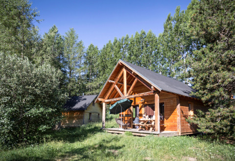 Gezellige houten chalet met overdekt terras en mensen aan tafel, tussen bomen bij Huttopia Font-Romeu, Frankrijk.