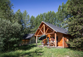 Gezellige houten chalet met overdekt terras en mensen aan tafel, tussen bomen bij Huttopia Font-Romeu, Frankrijk.