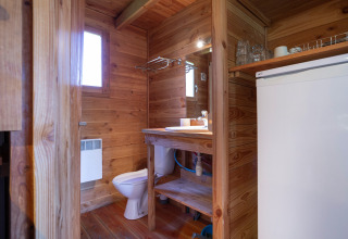 Interior de baño de cabaña con paredes de madera, inodoro, lavabo, espejo y frigorífico en Huttopia Font-Romeu.
