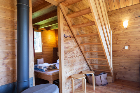 Binnenzicht van chalet ‘The Cabin’ met houten trap, kachel en bed bij Huttopia Font-Romeu in Frankrijk.