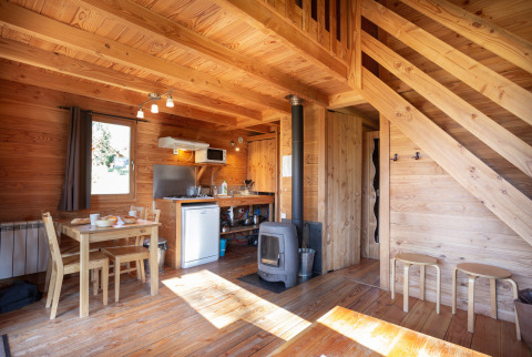 Wooden interior of Chalet 'The Cabin' at Huttopia Font-Romeu, France, with dining table and wood stove.
