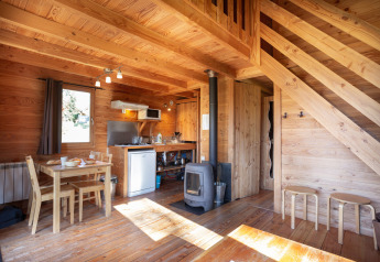 Wooden interior of Chalet 'The Cabin' at Huttopia Font-Romeu, France, with dining table and wood stove.
