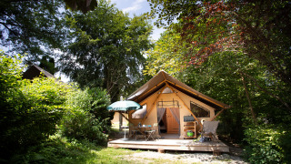 Tienda safari llamada Tente Trappeur con terraza, mesa, sillas y sombrilla, rodeada de naturaleza verde.