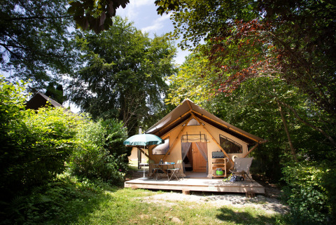 Tienda safari llamada Tente Trappeur con terraza, mesa, sillas y sombrilla, rodeada de naturaleza verde.