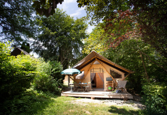 Tienda safari llamada Tente Trappeur con terraza, mesa, sillas y sombrilla, rodeada de naturaleza verde.