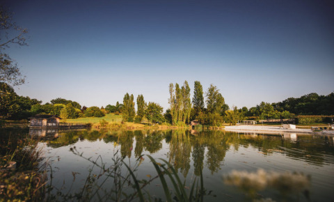 Vista panoramica di un lago tranquillo con alberi e prati a Huttopia Vallée du Lot, Nouvelle-Aquitaine, Francia.