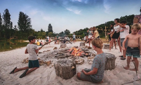 Børn og voksne hygger sig omkring et lejrbål på stranden i Huttopia Vallée du Lot, Frankrig.
