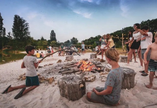 Børn og voksne hygger sig omkring et lejrbål på stranden i Huttopia Vallée du Lot, Frankrig.